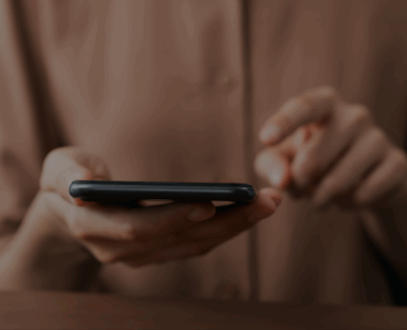 kobieta w beżowej koszuli, trzyma w dłoniach smartfon i przesuwa po nim palcem